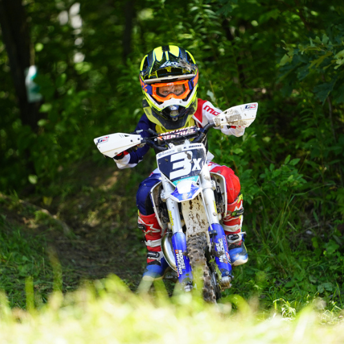Child riding a electric dirt bike through the trees, wearing a helmet and goggles.